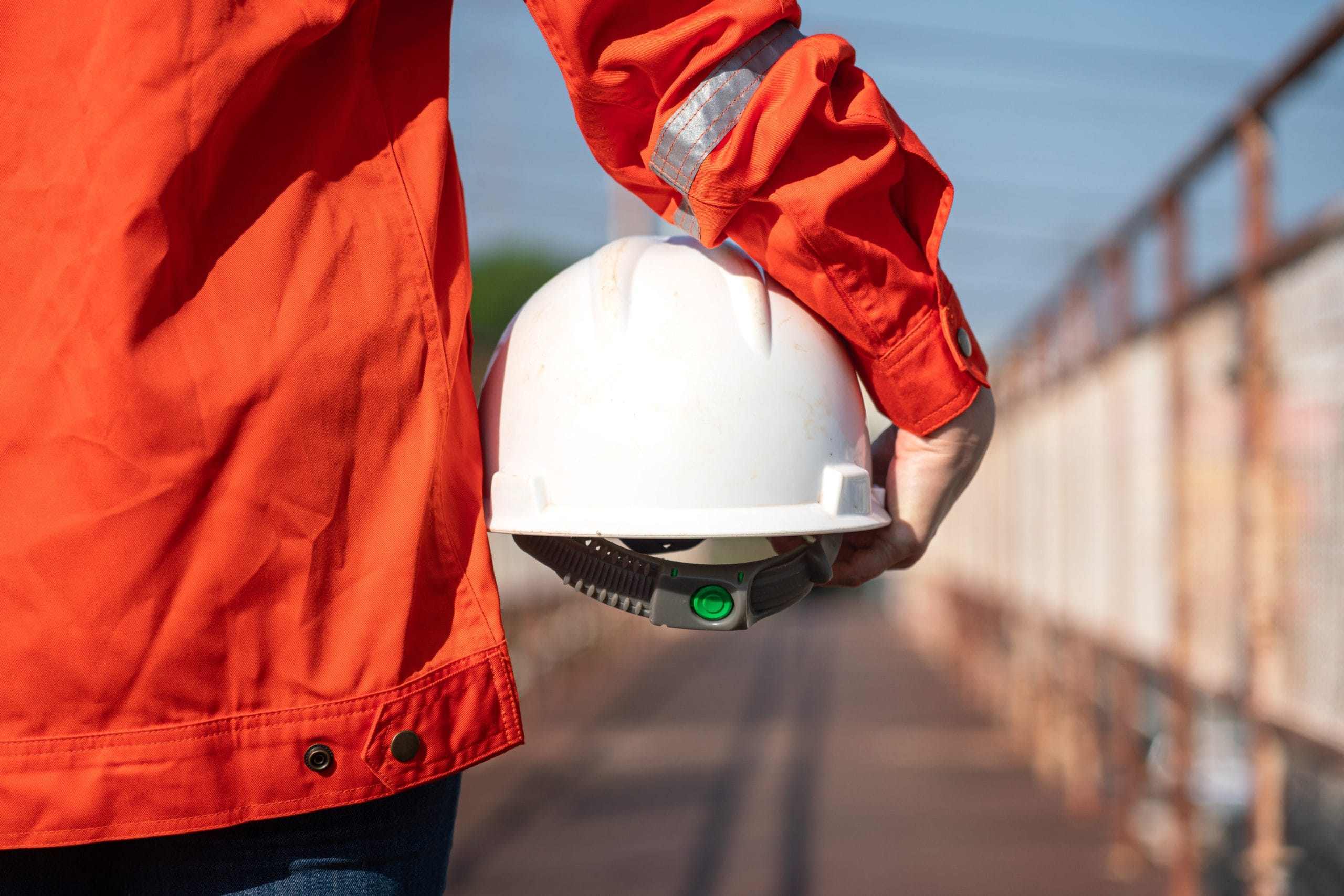 An engineer or construction worker is holding a white safety helmet, wearing orange coverall, standing on the working platform walkway.